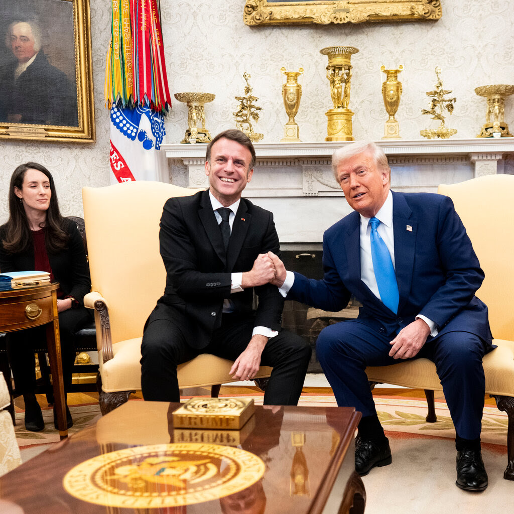 Two men in suits, one black and one navy, shake hands in yellow chairs in the Oval Office.