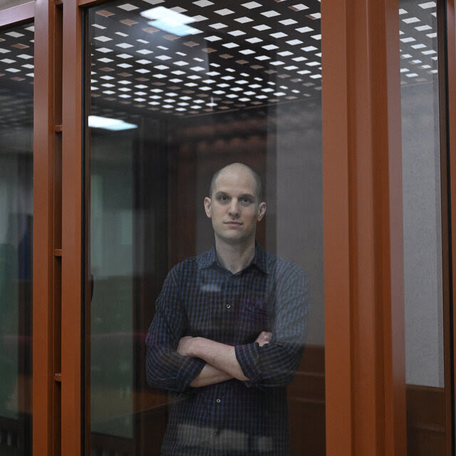 Evan Gershkovich, wearing a dark blue shirt and with his arms crossed, standing inside a glass cage. 