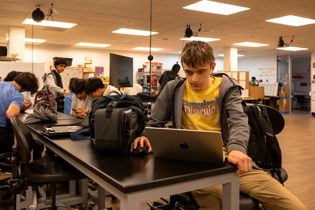 Students with backpacks sit at communal desks working on laptops.