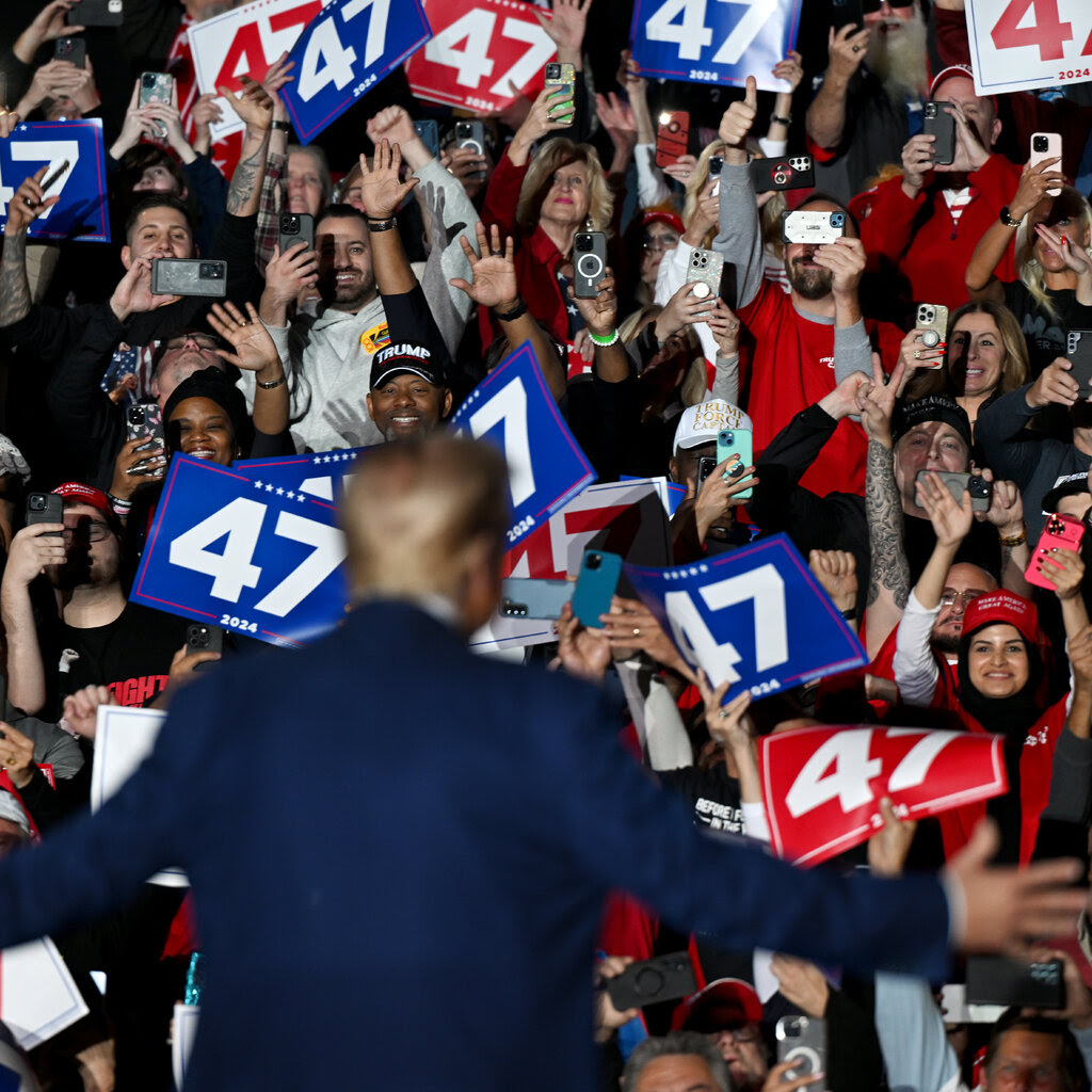 Donald J. Trump shown from behind with his arms stretch out before a large, cheering crowd. Several people in the crowd hold up signs reading “47.”