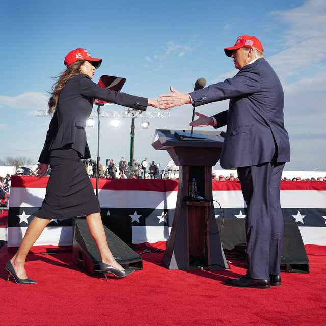Kristi Noem and Donald Trump, arms outstretched, greet each other at a political rally. Both wear red MAGA caps. Crowds are seen behind red, white and blue bunting.