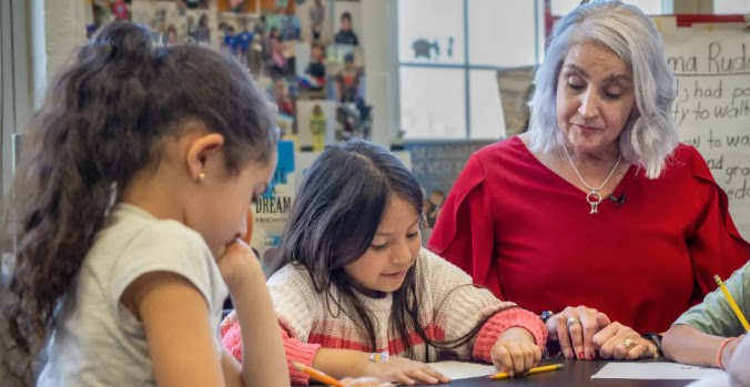 A teacher in a red shirt sitting at a table with children and teaching them.