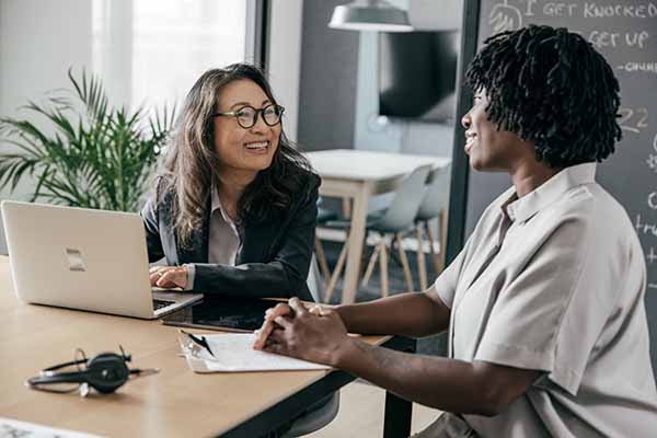 Two women speak to each other and work on laptop.