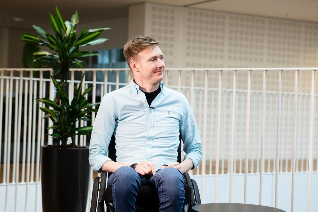 In a wheelchair, Jack Kavanagh wears a sky-blue shirt and blue jeans, smiling slightly to his left. Behind him is a potted dark-green plant, two sets of railings, and, behind them, an off-white wall.