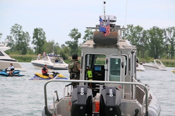 A male DNR conservation officer holds onto the side of a patrol boat moving among boats and personal watercrafts on a Michigan lake