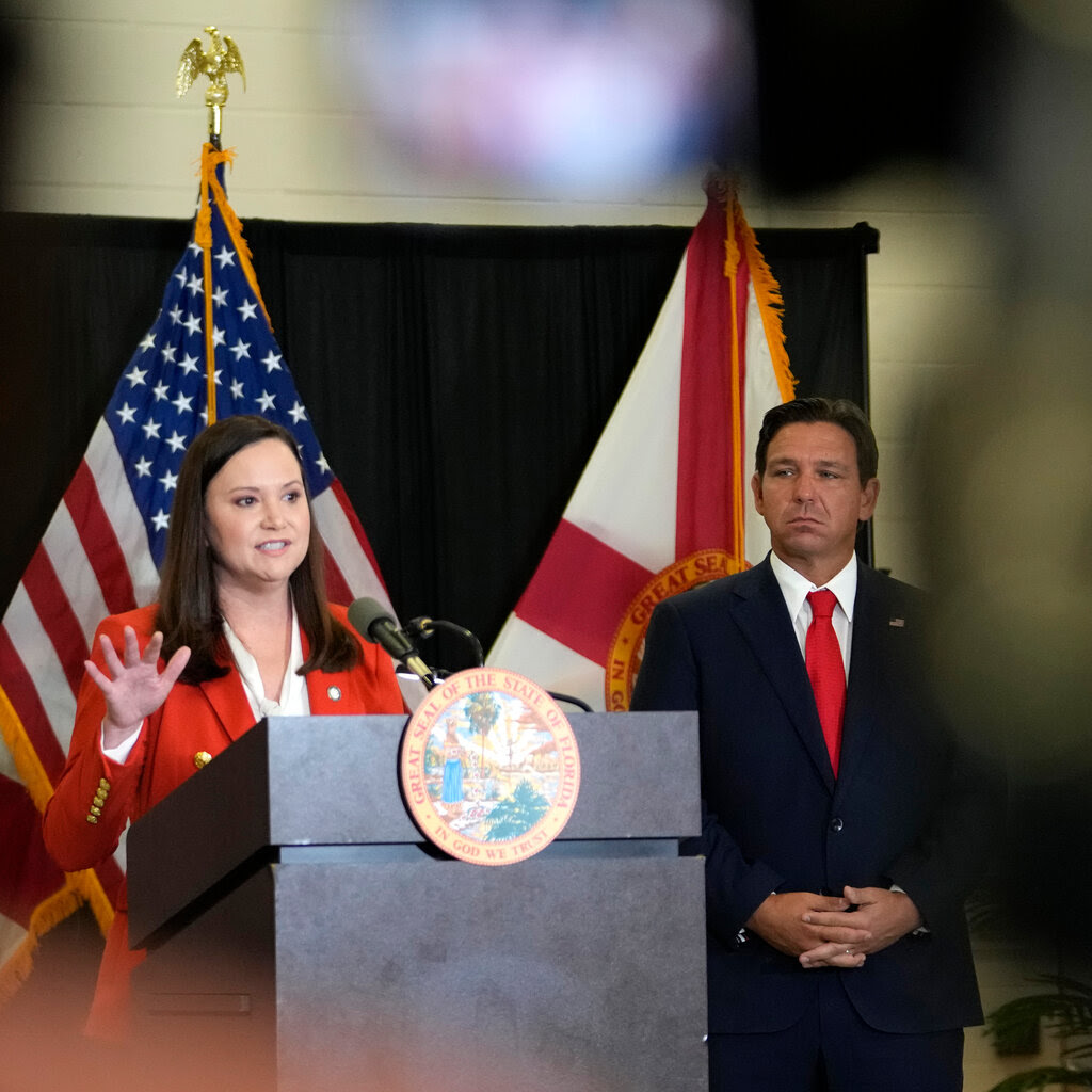 Gov. Ron DeSantis of Florida, wearing a dark suit and red tie, stands and listens as Florida Attorney General Ashley Moody speaks at a lectern. An American flag and a Florida state flag are displayed behind them.