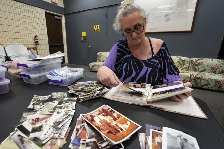 A woman sits at a table looking through a photo book. There are piles of photos on the table around the book. There are several plastic bins on the table beside her.