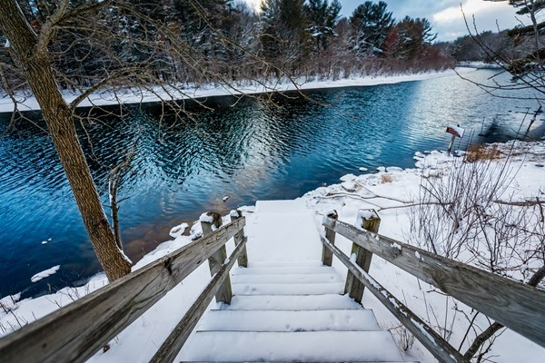 snow-covered wooden, railed steps go down to a calm, dark blue river, lined by dark green and auburn trees lightly snow-dusted