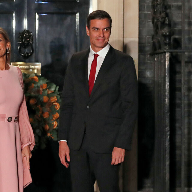 Pedro Sánchez, in a dark suit and red tie, stands in front of a door next to his wife, Begoña Gomez, who is wearing a pink dress with long sleeves.