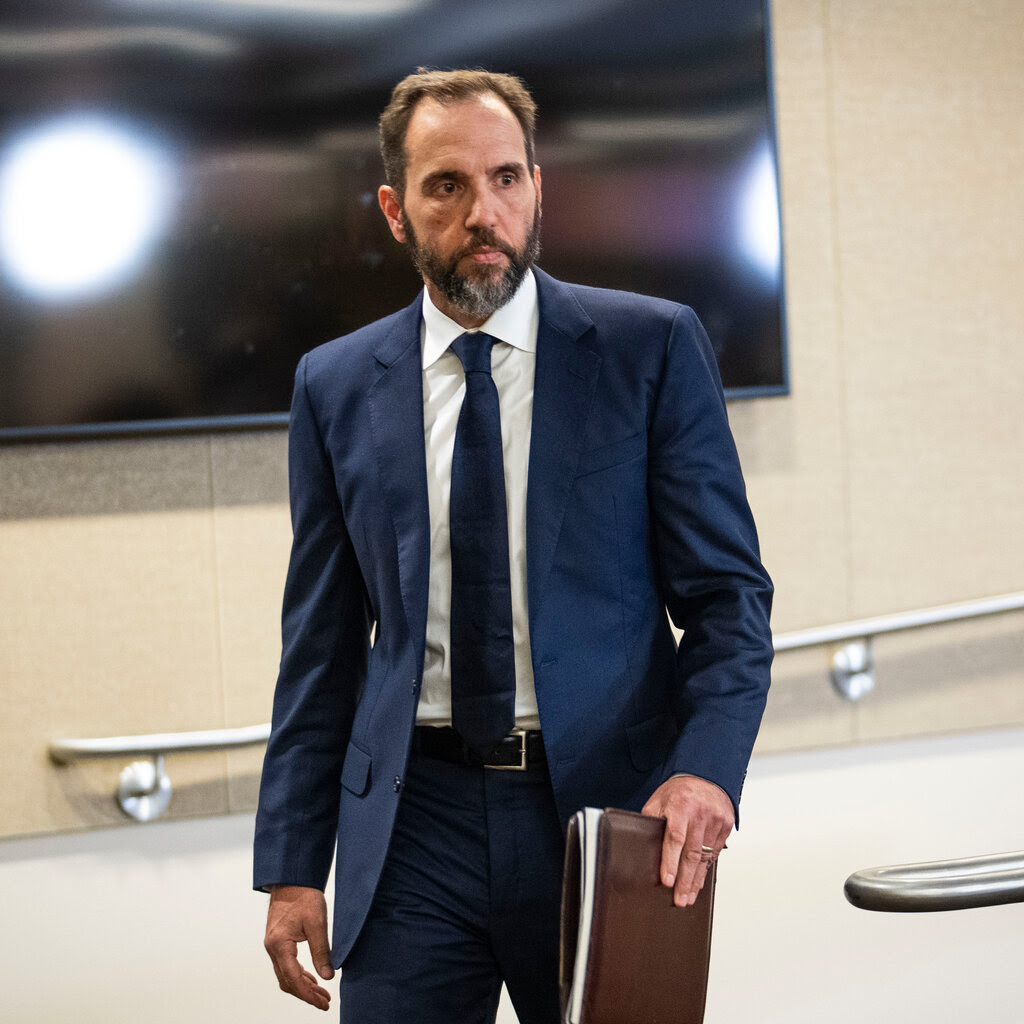 Jack Smith in a suit holds a brown leather folder.