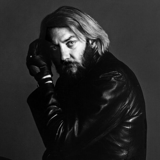 A man with long hair and a beard wearing gloves and a leather jacket looks directly at the camera.