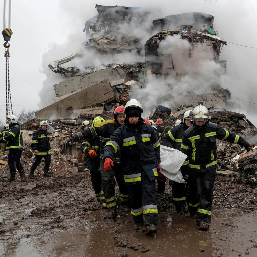 Rescue workers carrying a body in front of the smoking ruins of a destroyed building.