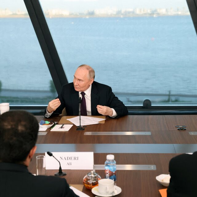 Vladimir V. Putin sitting at a table in front of a microphone with a wall of windows behind him.