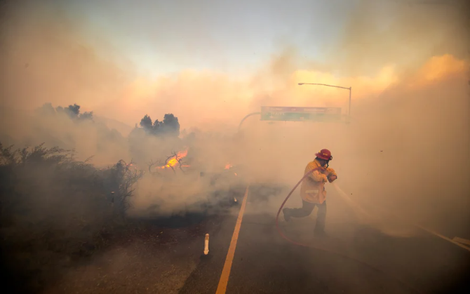 Photo shows a firefighter walking on a roadway while surrounded by thick gray and orange smoke as they carry and direct a hose at advancing fire in Irvine, California in October 2020