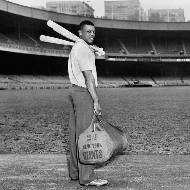 A black-and-white shot of a smiling Willie Mays in pants and a polo shirt, holding a duffle bag in his right hand and two baseball bats over his left shoulder. He is standing on the warning track in the outfield of Polo Grounds, the stadium in New York that hosted in the Giants.