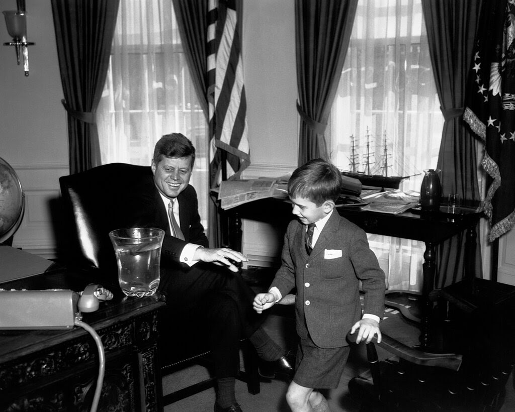 President John F. Kennedy Jr. sitting and smiling in the Oval Office as his young nephew Robert F. Kennedy Jr. looks at his desk and smiles. The boy is wearing a suit with shorts.