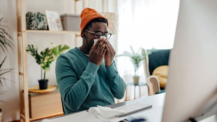 A man blows his nose as he sits at his desk.