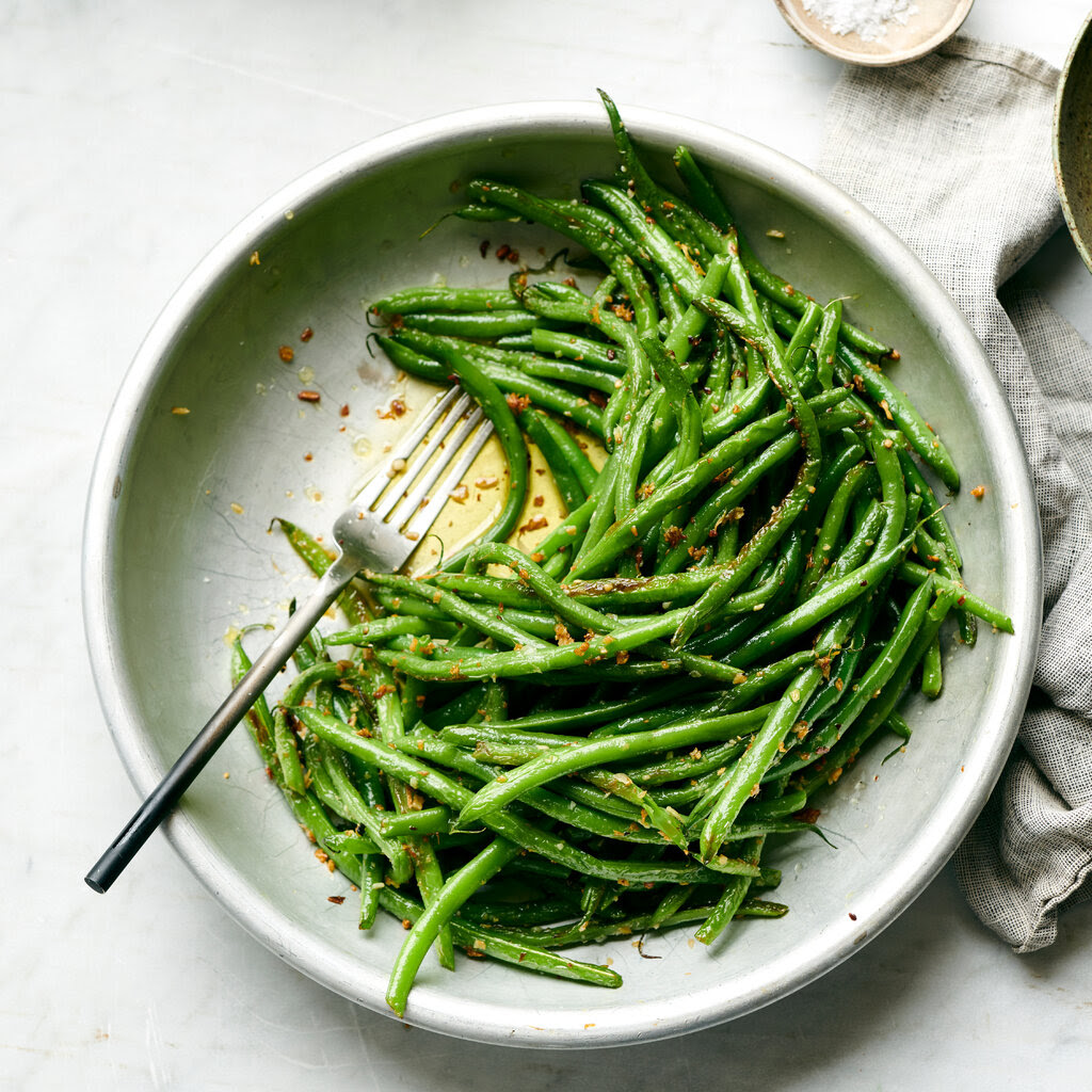 A bowl of green beans speckled with ginger and garlic is shot overhead. Just behind it is a textured gray napkin and a marbled background.