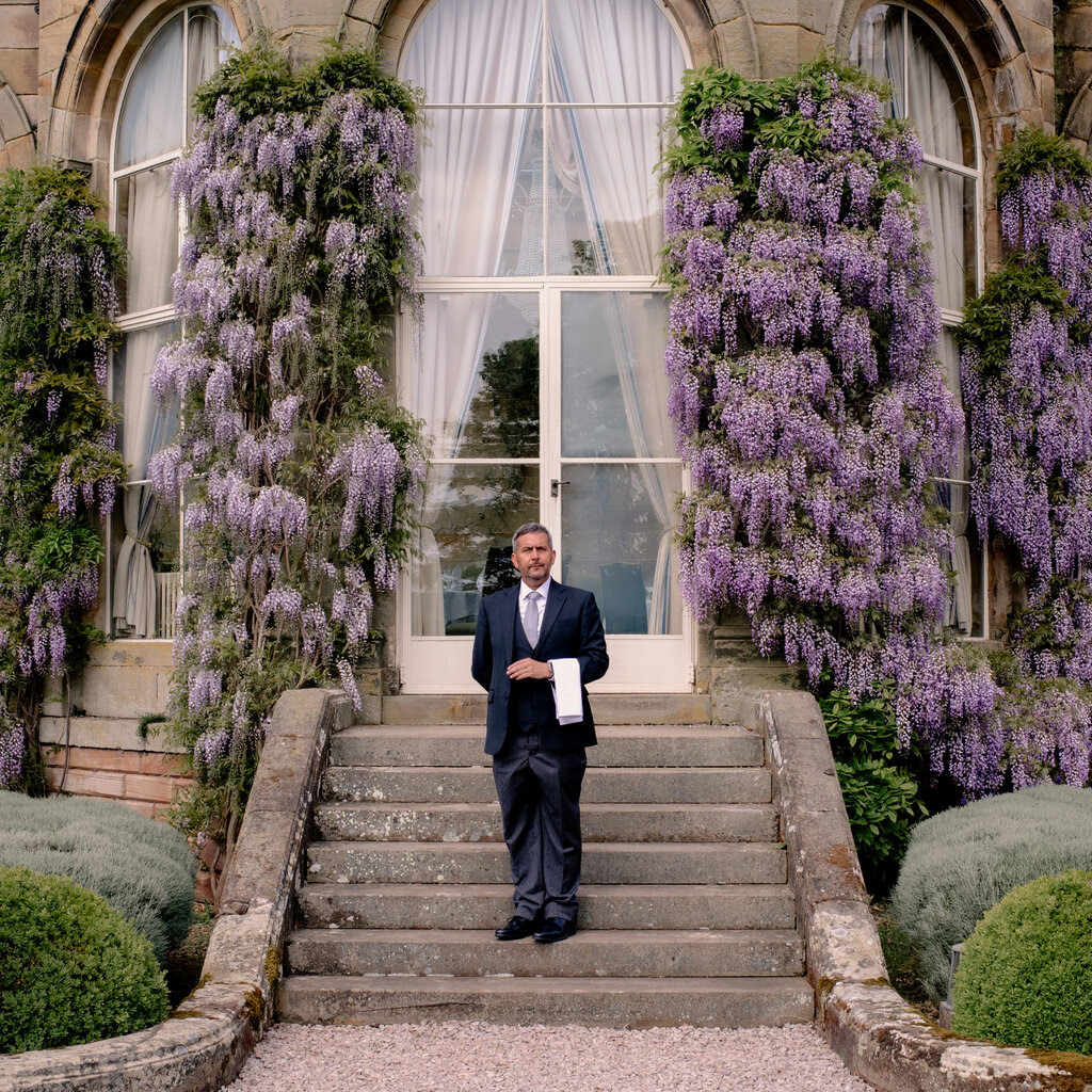 A butler stands outside a stately residence covered in wisteria. He has a white cloth draped over his arm.