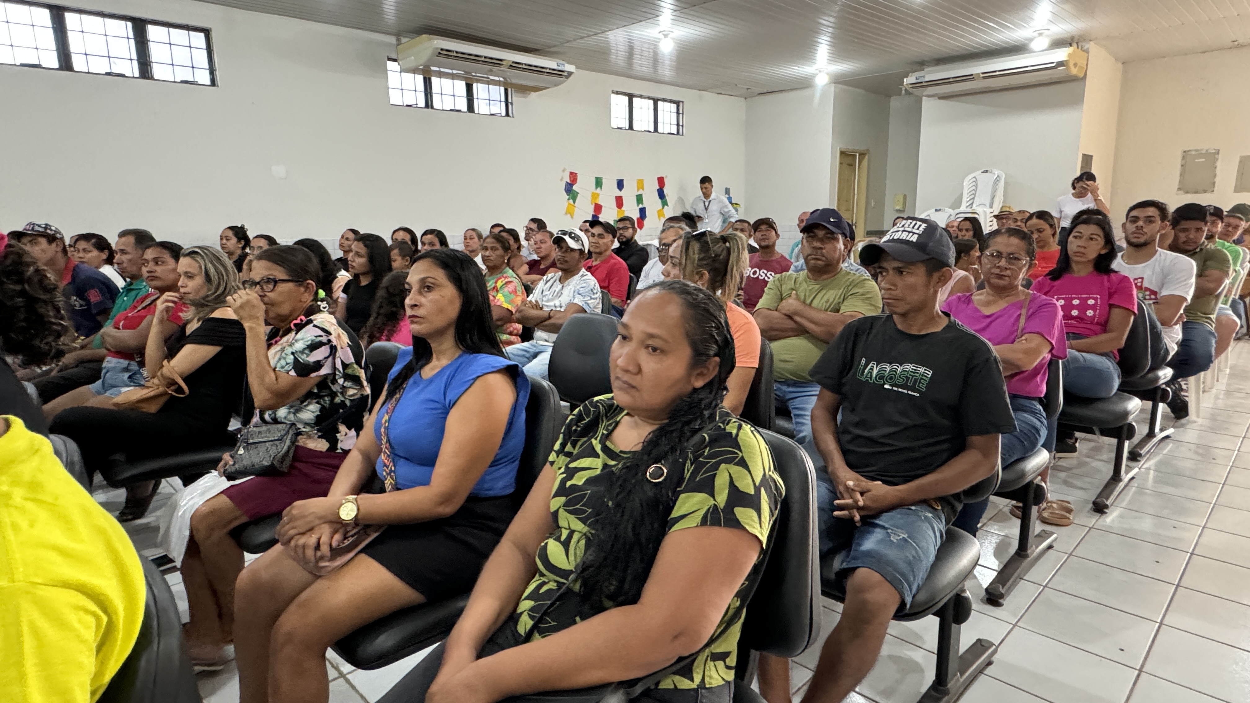 A imagem mostra um grupo de pessoas sentadas em fileiras dentro de uma sala, de frente para a frente, onde parece estar ocorrendo uma apresentação ou evento. A sala tem paredes brancas e é iluminada por luzes fluorescentes. Há decorações coloridas penduradas no teto, e uma pessoa está em pé na frente, perto de um quadro branco, o que sugere que isso poderia ser um encontro educacional ou comunitário. Os rostos de todas as pessoas foram obscurecidos com retângulos marrons para proteger sua privacidade.