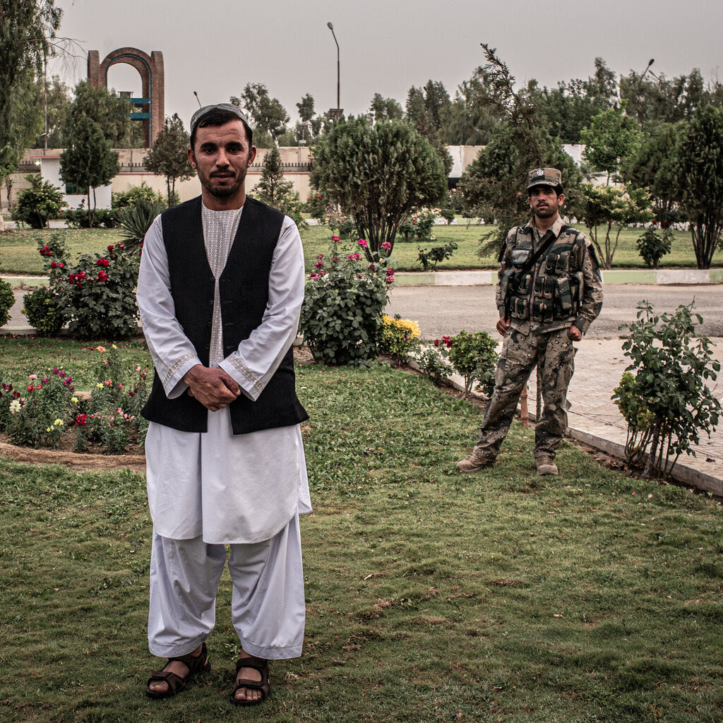 Abdul Raziq in traditional Afghan attire with his hands clasped at his waist, as a man in camouflage stands a few feet behind him.