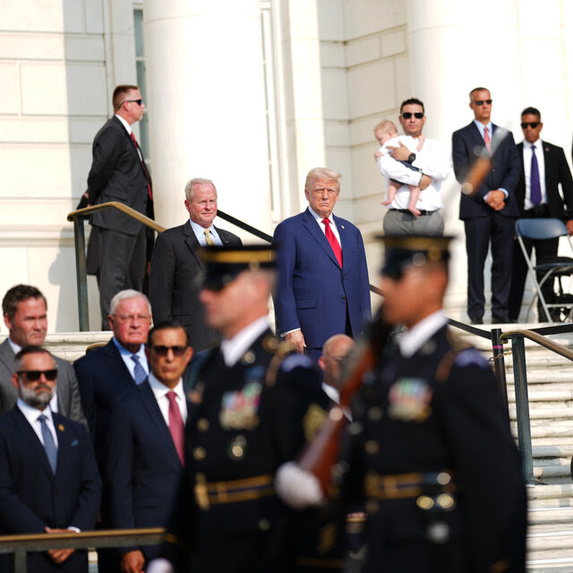 Donald Trump watches from a wide set of steps outside as uniformed soldiers carrying rifles march pass in procession.