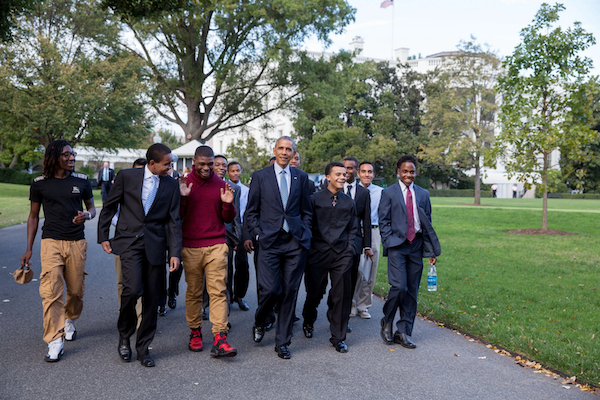 President Obama walking with a group of young men with medium to dark skin tones. They are smiling and talking with each other. 