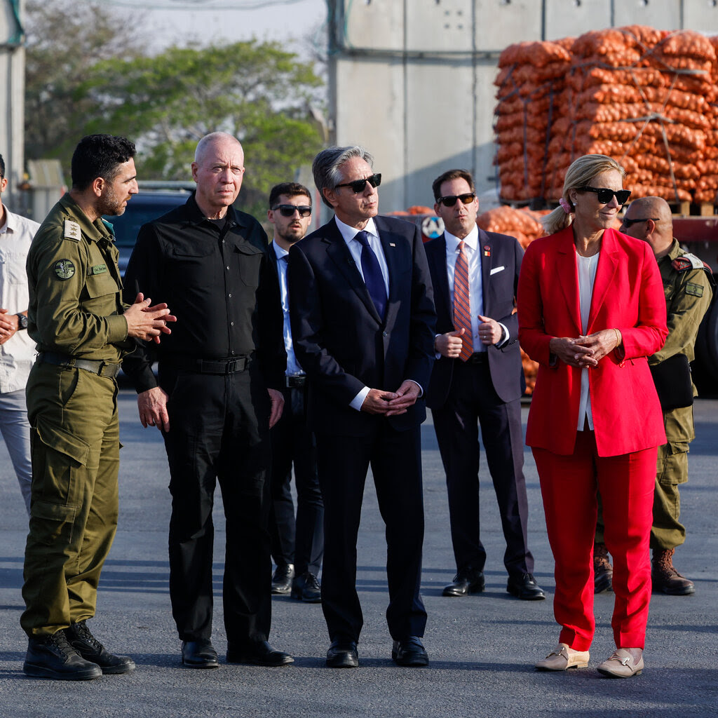 From left, a man in fatigues, a man dressed in all black, a man dressed in a suit and sunglasses, and a woman in a red pantsuit stand at a military checkpoint.