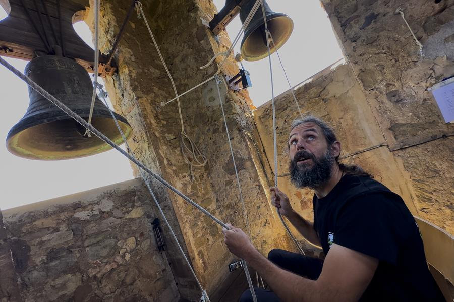 A man looks upwards while holding ropes attached to large metal church bells.