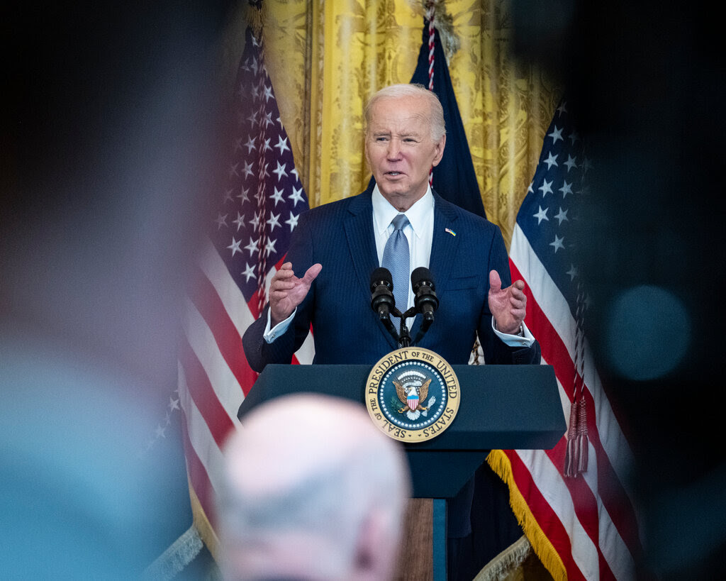 President Biden wearing a blue suit and tie standing at a podium in front of two American flags. 