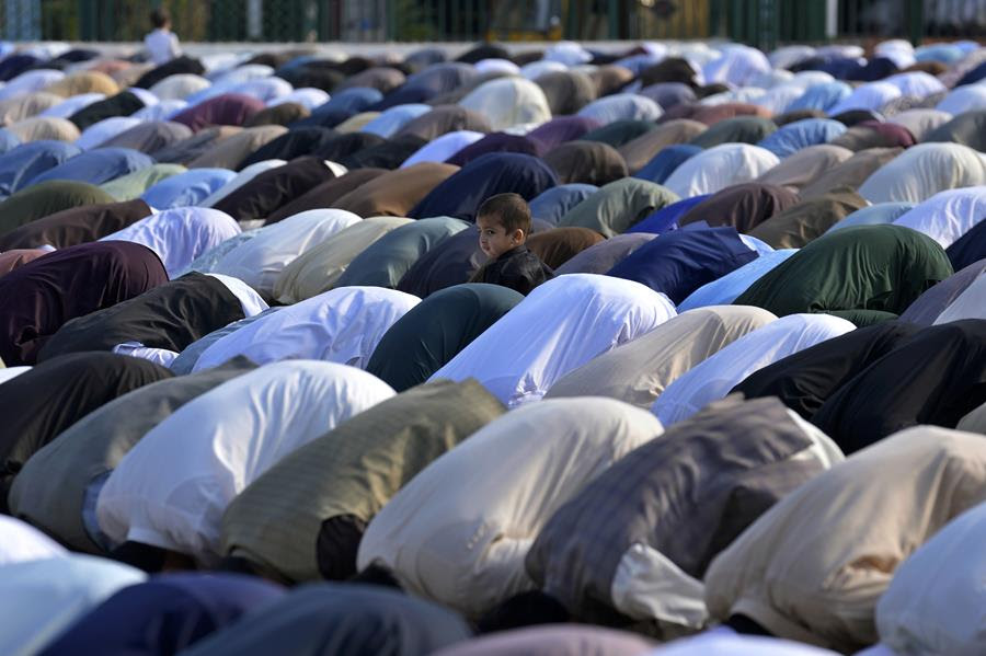 A group of Muslim people bow and perform an Eid al-Fitr prayer. A small child is standing amongst the crowd.