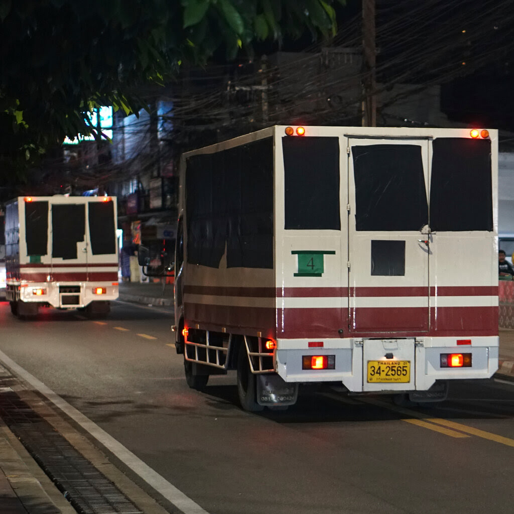 A handful of trucks, with their side and rear windows covered by black cloth, moving in a single file on a narrow urban road.