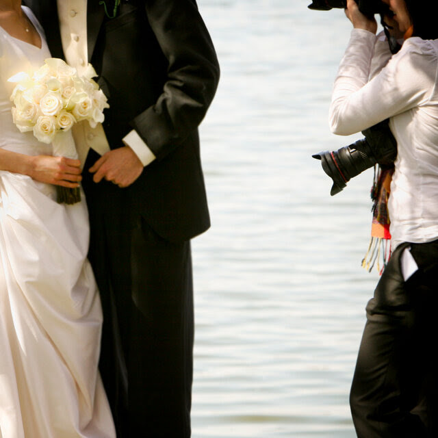 A bride and groom embrace on the left as a photographer takes photos. They are all standing in front of a body of water. 