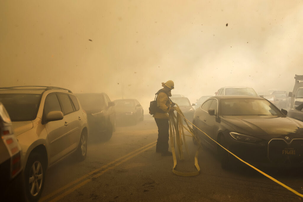 A firefighter stretches a line of yellow tape next to cars, with visibility obscured by smoke.