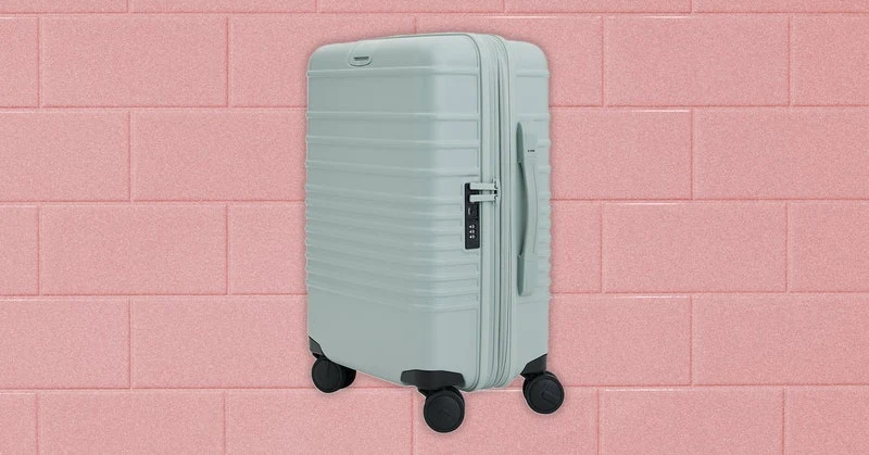 Small rectangular rolling luggage case with rounded corners and a long metal handle extended upward. Background: Pink rectangular tiles.