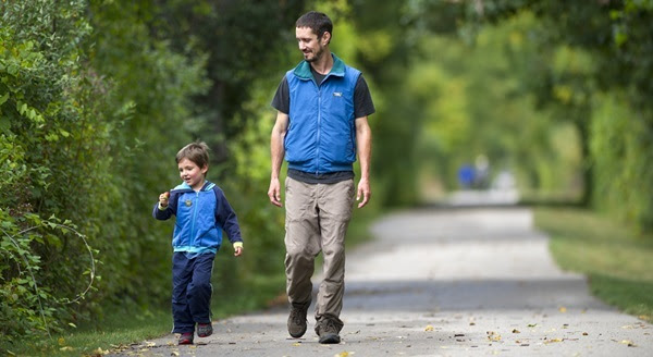 a father and young son, both dressed in jeans and light jackets, walk side by side on a paved trail, lined with green trees