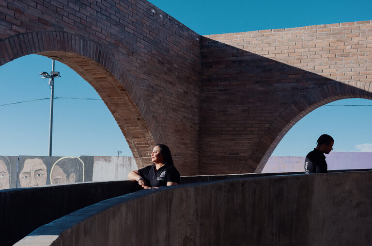 A woman stands facing the sun on a brick and concrete ramp. In the background is the border wall with colorful portraiture and mural painting. 