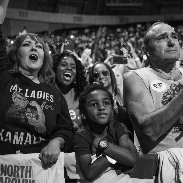 A black-and-white photo of a crowd of Kamala Harris supporters waving and cheering. 