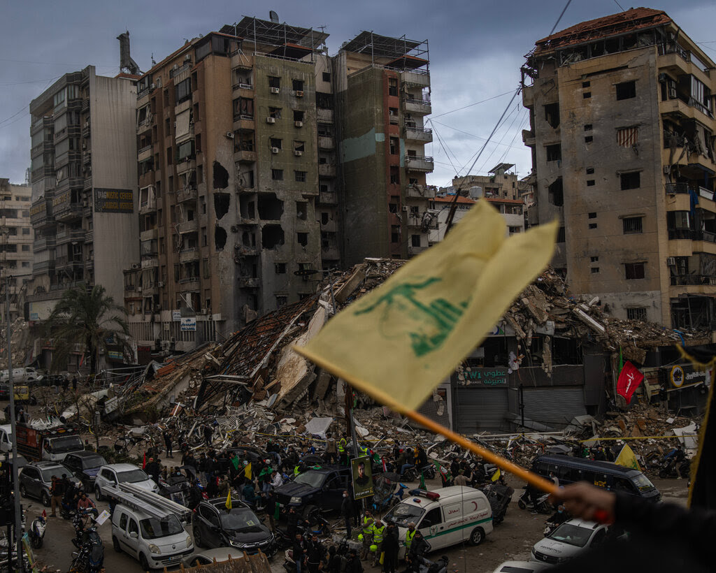 People on foot and in cars around a destroyed building in a city, with a yellow and green flag visible.