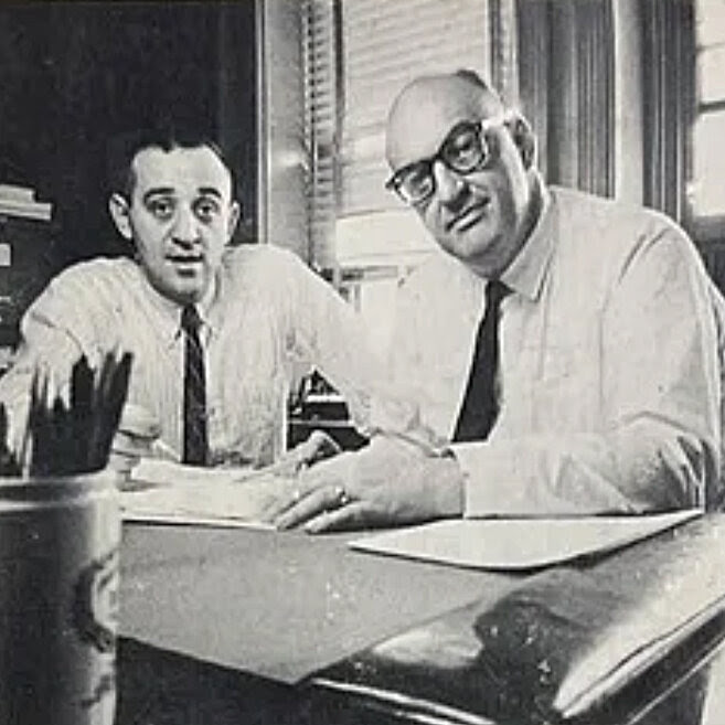 A black-and-white photo of two men in white shirts and black ties. 