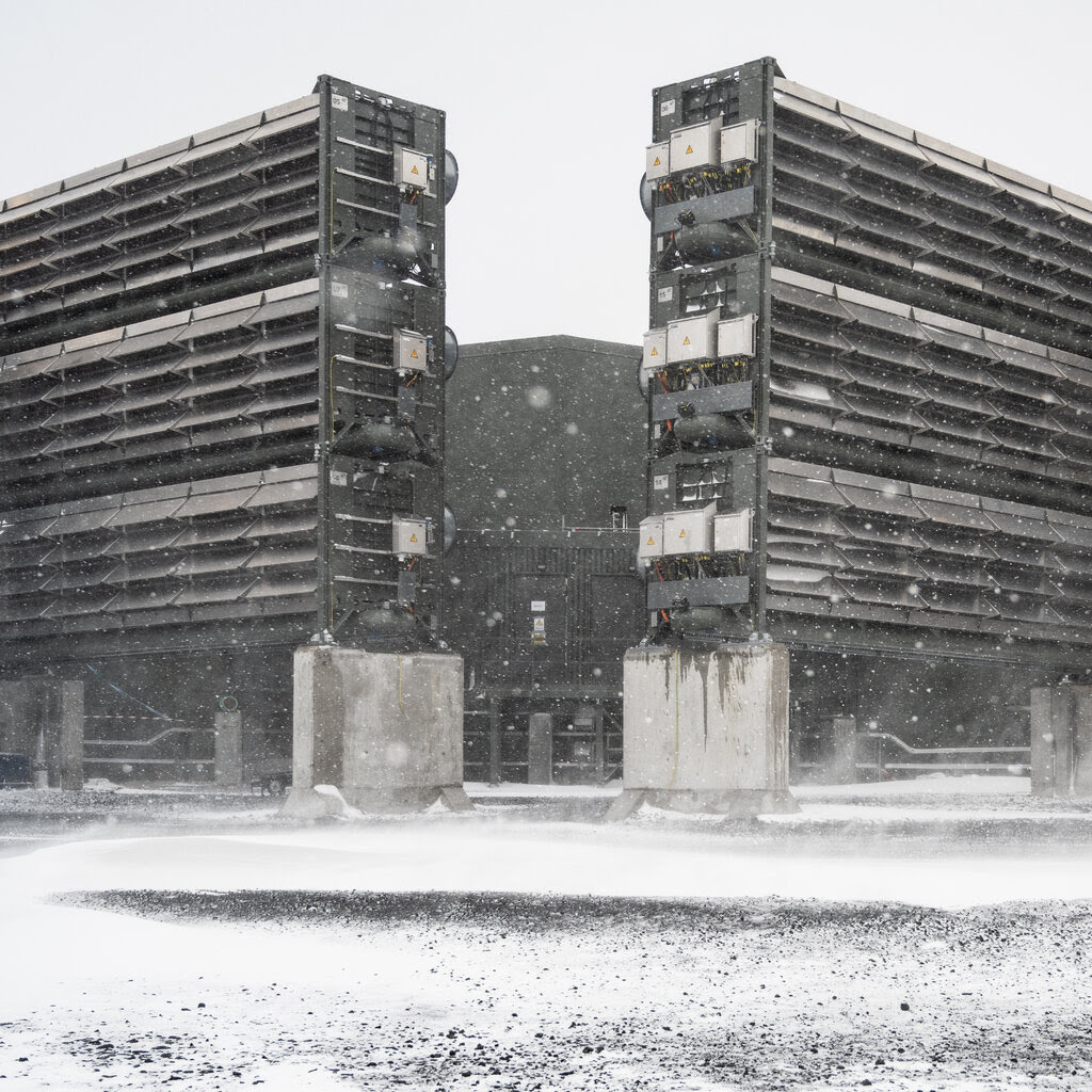 Amid swirls of snow, two tall, gray industrial structures, covered with louvers, stand at an angle to each other atop concrete pillars.