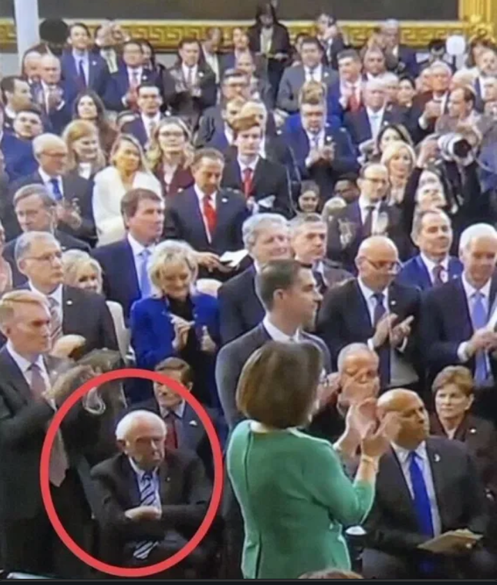 Image of applauding crowd with Bernie Sanders sitting and looking glum.