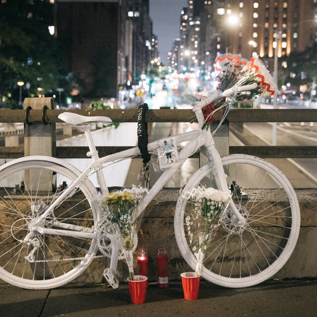 A bicycle that has been painted white is tied to railings near the scene of the accident where a man delivering food on a bicycle died in Manhattan. The lights of oncoming vehicles beam behind it.