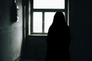 A very dark room is dimly lit by a single window in the corner. A woman is silhouetted against the window.