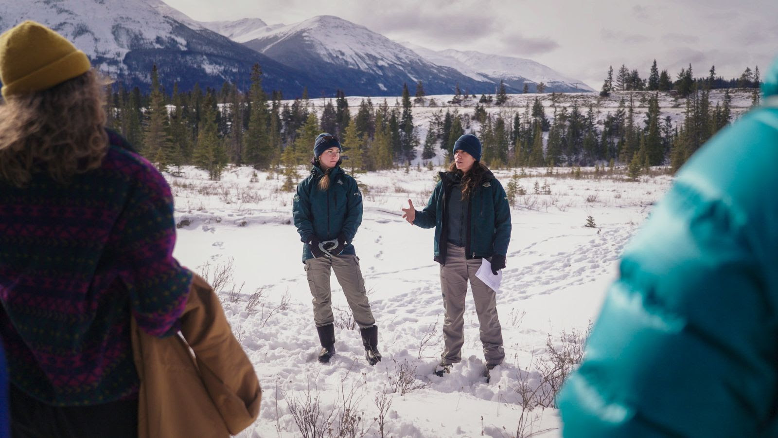 Two Parks Canada biologists are shown standing outside in the snow, with snowy mountains in the background, while presenting to a group of people.