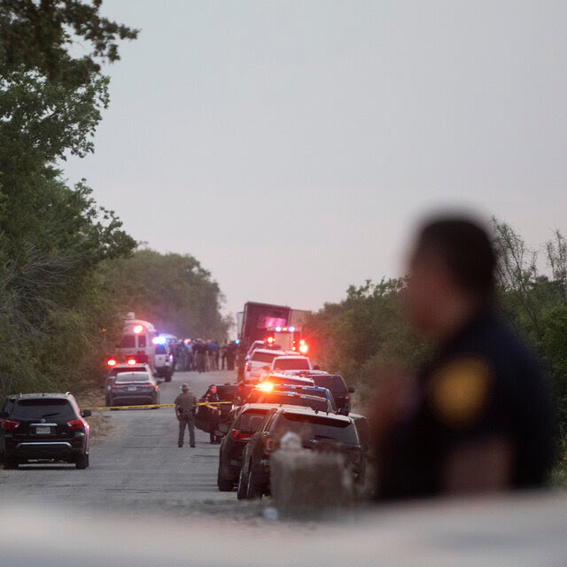 A road is lined with police and other vehicles, many with their lights flashing. Officers stand near a few of the vehicles. 