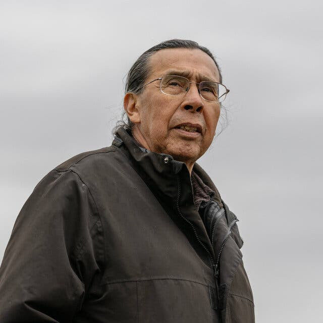 A man stands beneath an overcast sky, his hands inserted in the pockets of a tan coat.