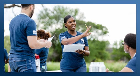 Woman in blue shirt and carrying white binder, giving instructions to other blue shirt CHWs in the field. Image has blue border around it.