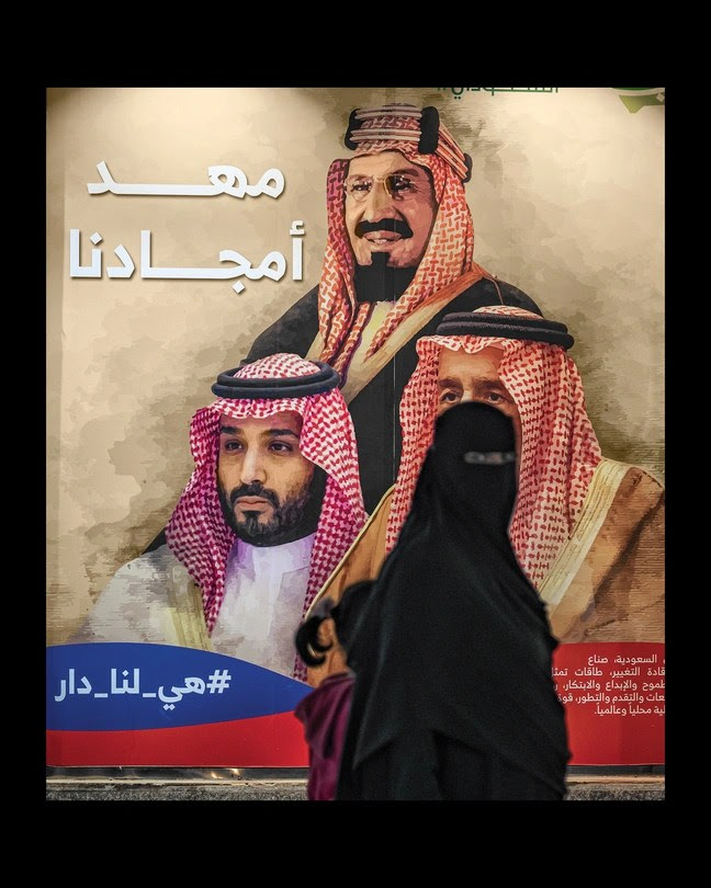 Une femme en niqab et abaya regarde la caméra tout en marchant avec un enfant devant une affiche plus grande que nature de la famille royale