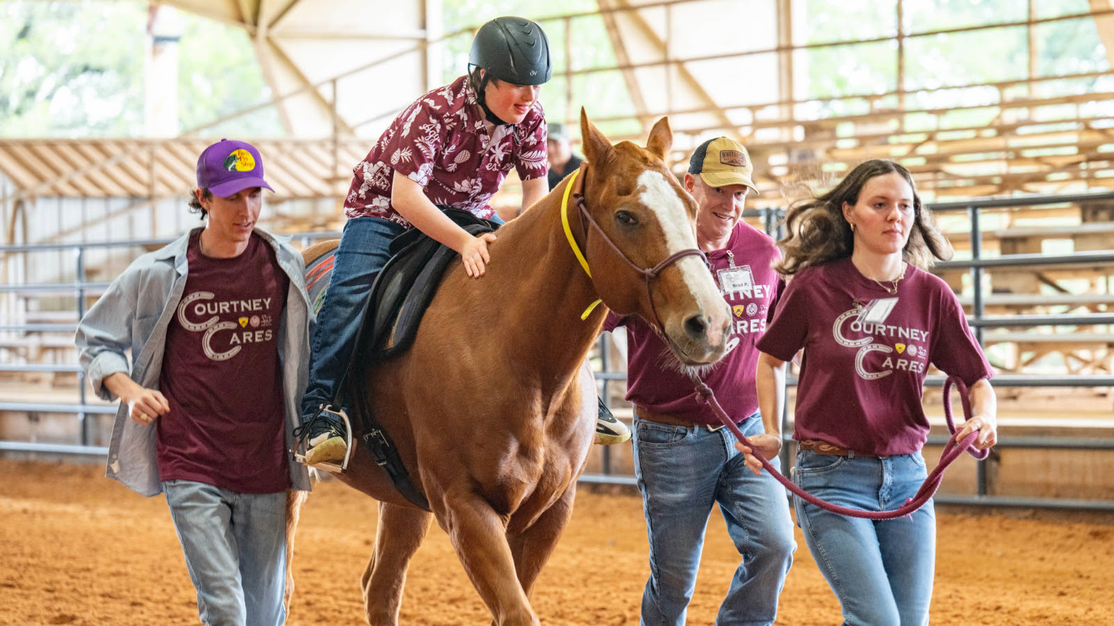 Three people wearing Courtney Cares shirts guiding a horse in an arena with a young man in a helmet riding